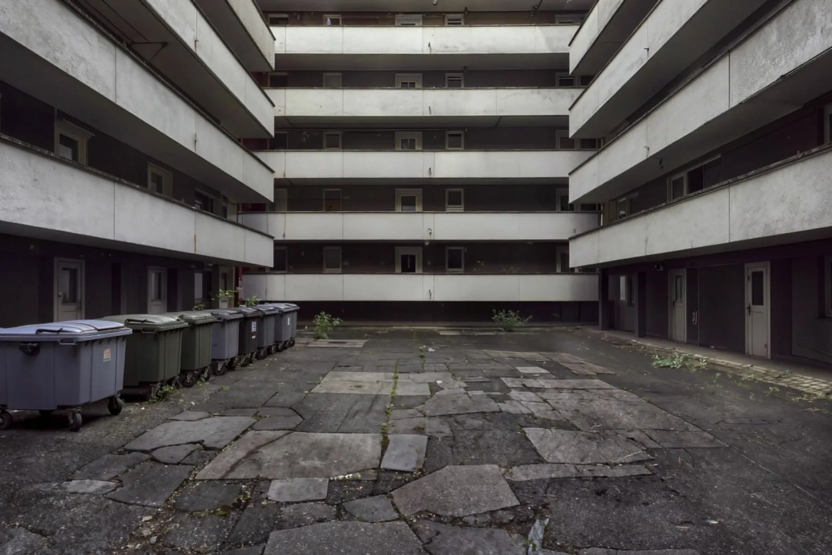 Grauer Innenhof einer Wohnanlage mit Laubengängen, rissigem Asphalt und Mülltonnen